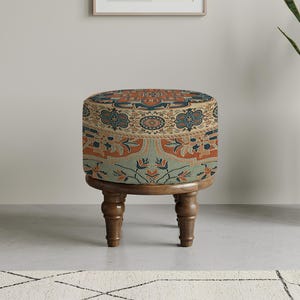 Op de afbeelding: Een kleine, ronde poef met een stoffen bovenkant met patronen in tinten van teal, oranje en beige. De poef heeft een houten basis met vier gedraaide poten. Het ontwerp doet denken aan een vintage tapijt. De poef is ongeveer 40 cm in diameter.
