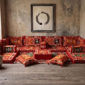 May include: A low-profile, red sectional sofa with patterned cushions and pillows. The sofa is arranged in a corner, with a matching ottoman and decorative pillows. A framed artwork hangs above the sofa, and a large vase sits nearby.