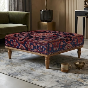 Pode incluir: Um otomano quadrado com estofamento de tecido azul-escuro e um padrão tradicional detalhado em tons de vermelho, rosa e creme. A peça é sustentada por quatro pernas de madeira de tom claro com um design torneado para estabilidade.