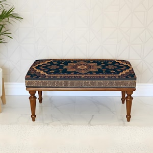 May include: A rectangular ottoman with a dark blue patterned top and brown wooden legs. The top features a detailed, ornate design in shades of gold, red, and brown. The legs are a rich, dark brown color, and the ottoman is placed on a white surface.