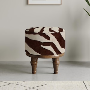 Puede incluir: Un otomán redondo con estampado de cebra en marrón y crema. La parte superior tapizada se asienta sobre una base de madera con cuatro patas torneadas. Esta pieza decorativa añade un toque de estilo safari a cualquier espacio habitable.