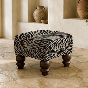 May include: A square ottoman featuring a black and white zebra print fabric upholstery. The piece is supported by four dark brown wooden legs with a rounded, turned design, providing a stable base for this low-profile furniture item.