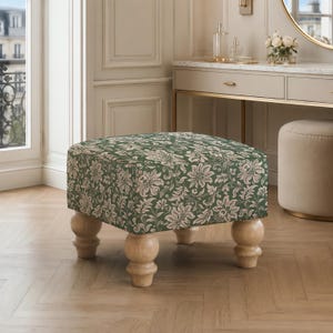 May include: A square ottoman featuring a dark green fabric upholstery with a repeating cream-colored floral and botanical pattern. The piece is supported by four light-colored, natural wood bun feet with a rounded, turned design at the base.