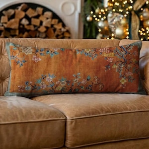 Può includere: Cuscino decorativo rettangolare con sfondo arancione bruciato e design floreale. Il cuscino presenta un motivo dettagliato di fiori e foglie nelle tonalità del rosa, blu e giallo. I bordi sono rifiniti con tessuto verde. Il cuscino è appoggiato su un divano in pelle marrone.