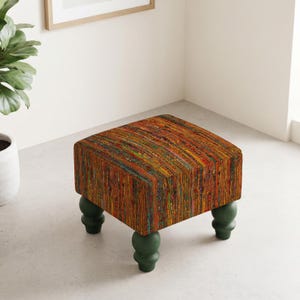 May include: A square ottoman featuring a multi-coloured, textured fabric upholstery with vertical stripes in shades of orange, red, yellow, and green. The piece is supported by four dark green, rounded, turned wooden legs in a classic style.