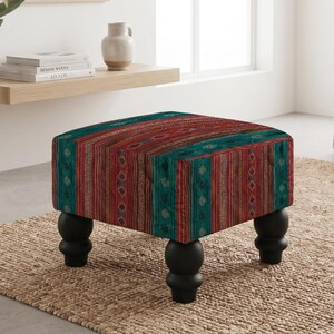 May include: A square ottoman featuring a vibrant fabric upholstery with vertical stripes in shades of deep red, teal, and brown, accented by small geometric patterns. The piece is supported by four rounded, black wooden bun feet.
