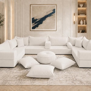 May include: A large white modular sectional sofa with a corduroy texture, featuring multiple back cushions, square armrests, and a matching round ottoman. Several square throw pillows are arranged on the floor in front of the seating area.