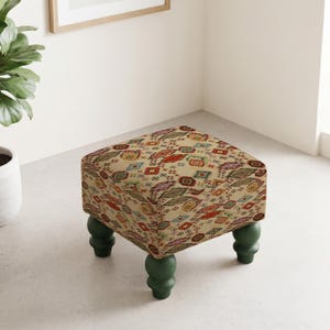 May include: A square ottoman featuring a cream-colored fabric with a multicolored geometric pattern in shades of red, blue, green, and purple. The piece is supported by four dark green, rounded, turned wooden legs that provide a stable base.