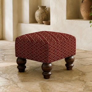 May include: A square ottoman featuring a deep red fabric upholstery with a repeating white geometric lattice pattern. The piece is supported by four dark brown, rounded wooden bun feet that provide a stable base for the cushioned seat.