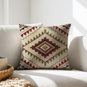 May include: Square throw pillow with a geometric pattern in cream, red, black, and brown. The design features a central diamond shape with smaller geometric elements and zig-zag borders. The pillow is placed on a white sofa.