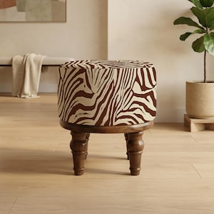 Puede incluir: Un otomán redondo y tapizado con estampado de cebra en marrón y crema. El otomán se asienta sobre una base de madera marrón oscuro con cuatro patas torneadas. Una pieza decorativa para una sala de estar o dormitorio.