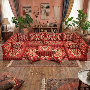 May include: A red sectional sofa with a geometric pattern, featuring multiple cushions and a low profile. The sofa is arranged on a patterned rug, creating a cozy and inviting seating area. The room is decorated with plants and decorative wall hangings.