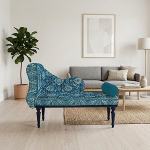 May include: A blue chaise lounge with a patterned design, featuring a curved back and dark blue legs. A teal bolster pillow rests on the seat. The lounge is placed in a living room setting with a gray sofa and a potted plant.