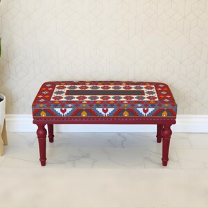 May include: A rectangular ottoman with a patterned top and red legs. The top features a geometric design in red, blue, yellow, and white. The legs are a deep red colour, and the ottoman is placed on a white floor.