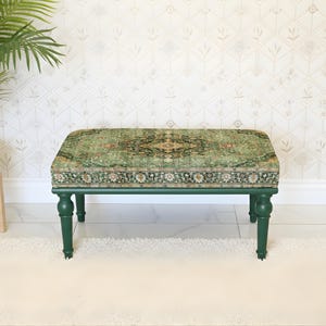 May include: An upholstered ottoman with a green, patterned rug design. The ottoman has dark green, turned legs and a rectangular shape. A potted plant and a white floor lamp are in the background.