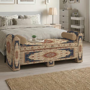May include: A rectangular storage bench featuring a traditional Persian-style rug pattern in shades of deep blue, cream, and red. The bench has two cylindrical beige bolster pillows at each end and a flat, upholstered top surface.