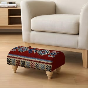 May include: A small footstool with a red patterned top, featuring blue, white, and black geometric designs. It has four short, light-wood legs. The footstool is placed in front of a cream armchair, adding a touch of colour.