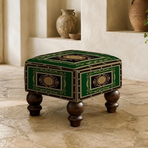Könnte beinhalten: Ein quadratischer Hocker mit tiefgrünem Stoffbezug, schwarzen rechteckigen Paneelen, goldenen kreisförmigen Medaillons und komplexen cremefarbenen Randmustern. Das Möbelstück steht auf vier dunkelbraunen, abgerundeten Holzfüßen.