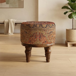 Op de afbeelding: Een ronde poef met een patroon, tapijtachtig bovenblad in tinten rood, bruin en blauw. De poef staat op vier donkere, gedraaide houten poten. Het stuk is een decoratief meubelstuk.