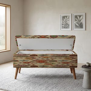 Può includere: Una panca contenitore rettangolare con tessuto di base beige caratterizzato da un motivo geometrico multicolore in rosso, verde e blu. La panca presenta quattro gambe affusolate in legno e un coperchio incernierato che rivela un vano interno bianco.