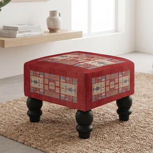May include: A square ottoman featuring a vibrant red fabric base with a repeating rectangular pattern of intricate floral and geometric motifs in shades of blue, cream, and gold. The piece is supported by four rounded, black bun-style feet.
