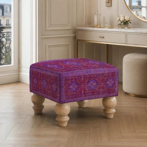 May include: A square ottoman featuring a vibrant purple and red patterned fabric with intricate floral and geometric motifs. The piece is supported by four light-colored, rounded wooden bun feet, providing a stable base for the upholstered seat.