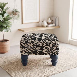 May include: A rectangular ottoman featuring a black and beige abstract floral pattern on its upholstered top. The piece is supported by four dark blue, rounded, turned wooden legs that provide a stable base for the cushioned seat.