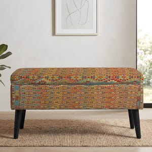 May include: A rectangular storage bench featuring a vibrant, multi-colored woven fabric with a complex geometric pattern in shades of red, orange, yellow, and teal. The bench is supported by four tapered black legs and has a hinged top lid.