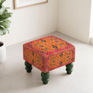 May include: A square ottoman featuring a vibrant orange fabric with a multi-colored floral and geometric pattern. The piece is supported by four dark green, rounded, bulbous wooden legs that provide a sturdy base for the upholstered seat.