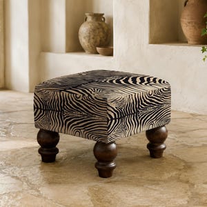 May include: A square ottoman featuring a black and beige zebra stripe pattern on the upholstered top and sides. The piece is supported by four dark brown, rounded wooden bun feet that provide a stable base for the cushioned seat structure.