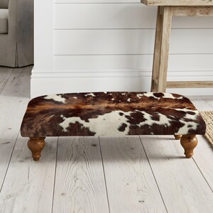 Può includere: Un poggiapiedi rettangolare basso rivestito con un motivo in pelle bovina marrone e bianca. L'oggetto presenta una superficie superiore piatta con una texture naturale di pelle animale ed è sostenuto da quattro gambe in legno corte e arrotondate.