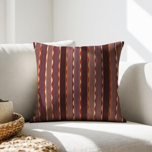 Può includere: Cuscino decorativo quadrato con un motivo a righe verticali nei toni del bordeaux, viola e oro. Il cuscino è su un divano bianco. Le strisce hanno un design ondulato.