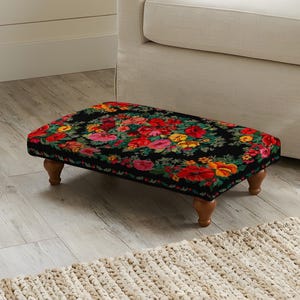 May include: A rectangular footstool featuring a black fabric top with a vibrant floral pattern in shades of red, orange, yellow, and green. The footstool is supported by four short, rounded, light brown wooden legs.