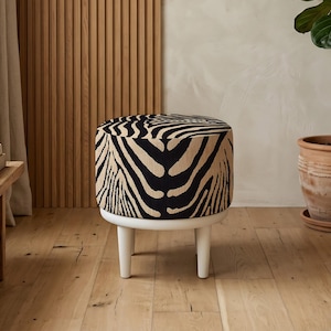 Könnte beinhalten: Ein runder Hocker mit einem Stoff mit Zebra-Print in Schwarz und Beige. Der Hocker steht auf einem weißen Sockel mit drei kurzen, weißen Beinen. Der Hintergrund umfasst eine Holzwand und eine Topfpflanze.