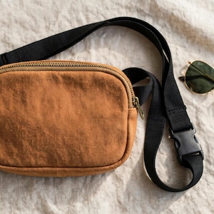 May include: A tan canvas belt bag with a black adjustable nylon strap and a black plastic side-release buckle. The bag features a top zipper closure with a brass-toned metal pull. Beside the bag are gold-rimmed aviator sunglasses.