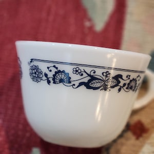 May include: White teacup with a blue floral design around the rim. The cup has a handle and a smooth, glossy finish. The floral pattern includes flowers and leaves.