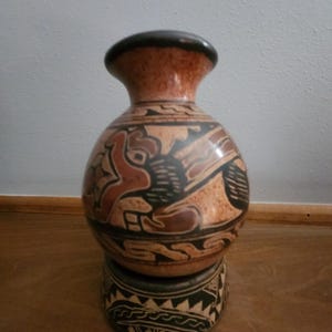 Peut inclure: Un petit vase rond en céramique brune avec des motifs géométriques noirs et bruns. Le vase est sur un petit piédestal rond en céramique noir et blanc.