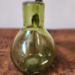 May include: A small, round, green glass bottle with a narrow neck. The bottle has a textured surface and appears to be antique.