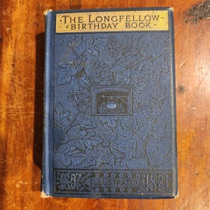 Puede incluir: Un libro de tapa dura azul titulado "The Longfellow Birthday Book" con letras doradas. La portada presenta un intrincado diseño floral y el texto "1897 Illustrated 1897".