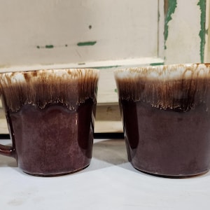 以下が含まれることがあります： ドリップ釉薬のデザインが施された2つの茶色のセラミックマグ。マグカップは、濃い茶色のベースカラーに、縁の周りに明るい茶色と白の釉薬が施されています。各マグカップにはハンドルが付いています。