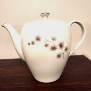 May include: White ceramic teapot with a lid and handle. The teapot features a floral design in brown and gray. The teapot is sitting on a dark brown wooden surface.