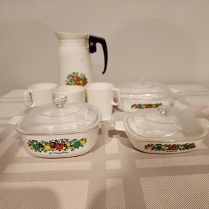 May include: A white vintage kitchenware set featuring a pitcher with a black handle, four mugs, and two covered casserole dishes. The dishes and pitcher are decorated with a colourful fruit and vegetable design. The casserole dishes have clear glass lids.
