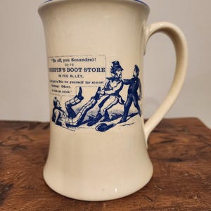 Könnte beinhalten: Eine weiße Keramiktasse mit blauem Rand und einer blauen und weißen Illustration einer Schuhladenwerbung. Der Text auf der Tasse lautet "Be off, you Scoundrel! Go to Crispin's Boot Store in Peg Alley, and get a Pair for yourself for almost Nothing: Officer.  Let him go quick."