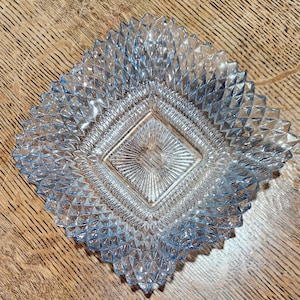Puede incluir: Un plato cuadrado de vidrio azul claro con un patrón tallado en diamante. El plato tiene un borde texturizado en relieve y un diseño central en forma de diamante. El vidrio es transparente, sobre una superficie de madera.