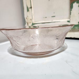 Puede incluir: Un cuenco de cristal rosa con un diseño floral. El cuenco tiene una forma ancha y poco profunda y se asienta sobre una pequeña base. El diseño floral está grabado en el cristal, creando una textura delicada. El cuenco es probablemente vintage o antiguo.