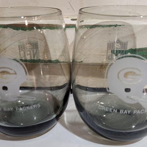 Könnte beinhalten: Zwei Weingläser ohne Stiel mit Grautönung. Jedes Glas zeigt ein weißes Helmdesign und die Worte "Green Bay Packers" in geschwungener Schrift. Das NFL-Logo ist schwach sichtbar.