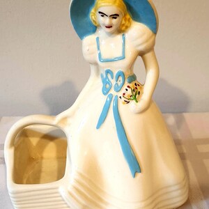 May include: A vintage ceramic figurine of a woman in a white dress and a blue hat. The figurine has a basket attached and is decorated with blue accents and a small bouquet of flowers. The woman's hair is blonde, and her lips are red.