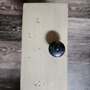 May include: A rectangular, off-white painted wooden block with a black knob attached. The wood grain is visible, and the block shows signs of wear. The knob is round with a screw in the center. The block is likely part of a larger piece of furniture.