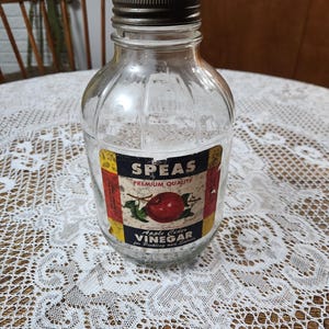 Op de afbeelding: Een heldere glazen fles met een zilveren deksel en een etiket met de tekst "Speas Premium Quality Apple Cider Vinegar for Pickling and Salads". Het etiket toont een rode appel met een groen blad.