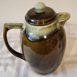 May include: A ceramic teapot with a dark brown glaze, accented with a lighter green and white trim. The teapot has a handle, a spout, and a lid with a small knob. The overall shape is rounded and the design is vintage.
